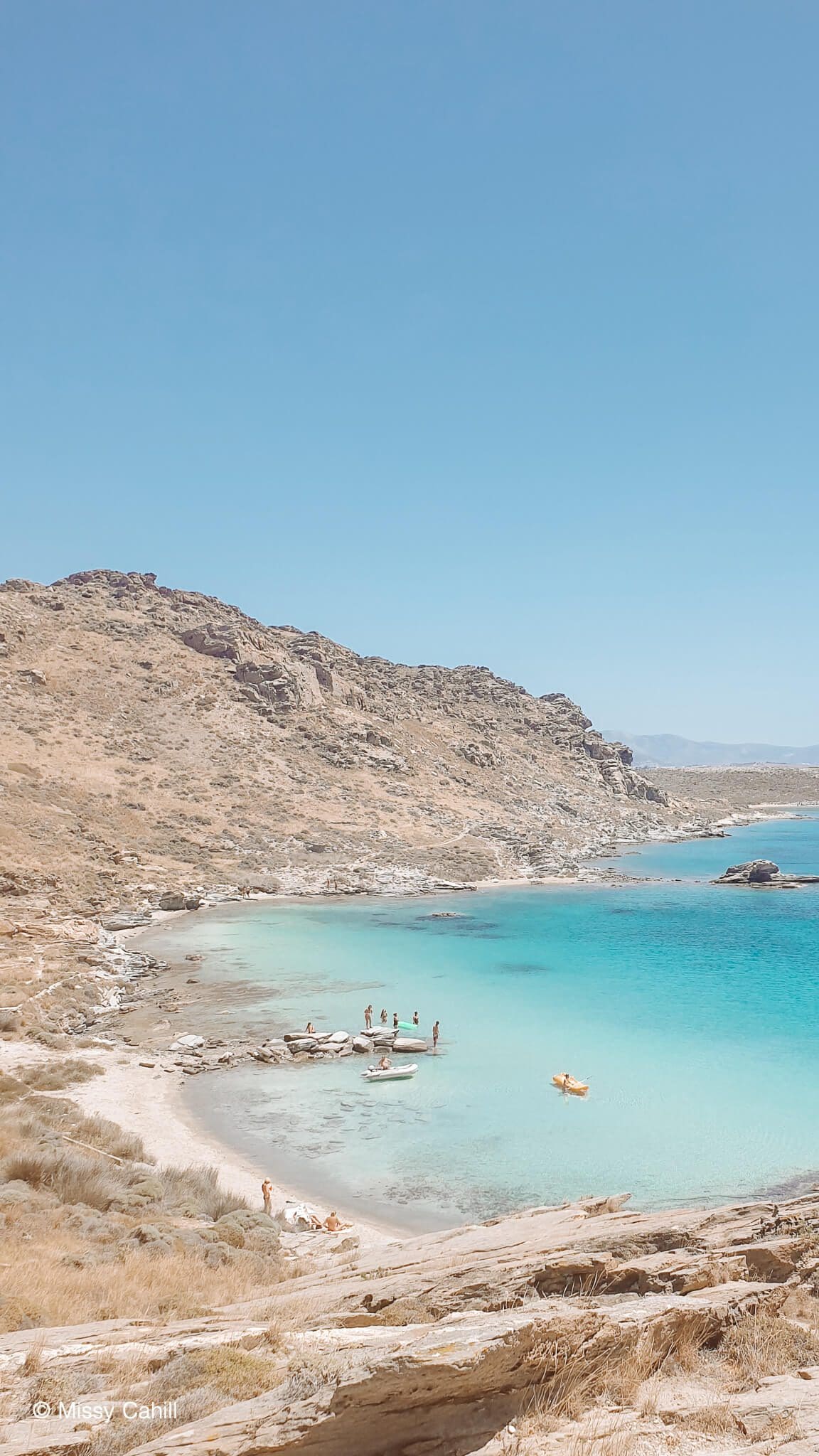 View of a secluded cove at Monastiri Beach Paros, the water is different shades of blue and the surrounding hills are full of scrub bush.