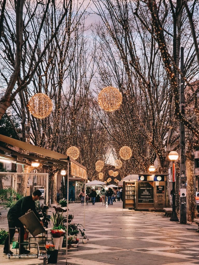 La Rambla in Palma de Mallorca is lit up at dusk with circular light fixtures hanging from trees. Lights have been strung up around tree trunks, and a flower vendor tidies up his stall.