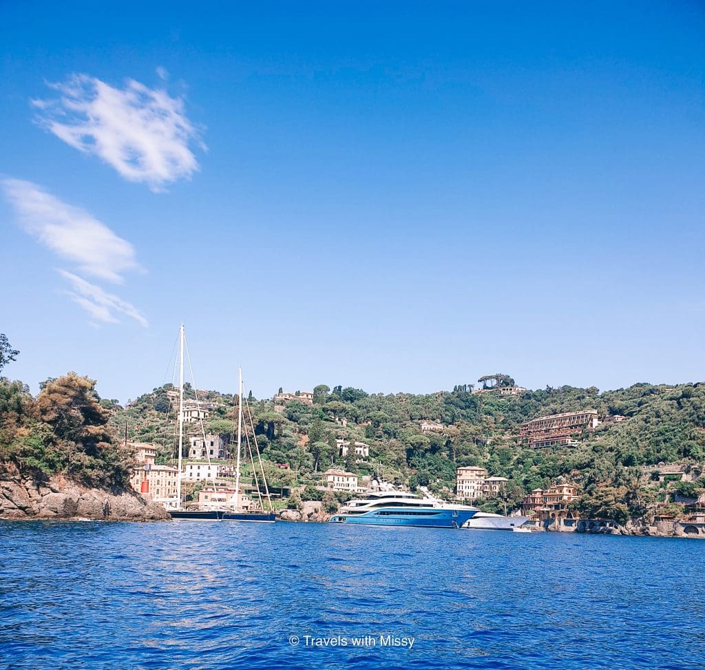 View of Portofino Bay with several large superyachts in the background, as well as rolling hills covered in trees and expensive homes.