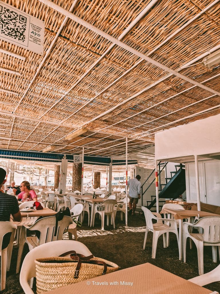 White plastic chairs of Ayo's Nerja sit in the sandy floor and a rattan ceiling overhead.