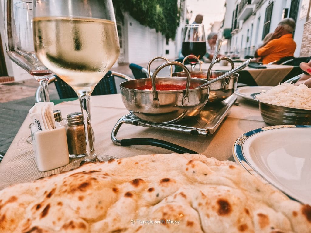View over a table with a naan bread, glass of wine and dishes containing Indian food in the foreground in A Taste of India Nerja.
