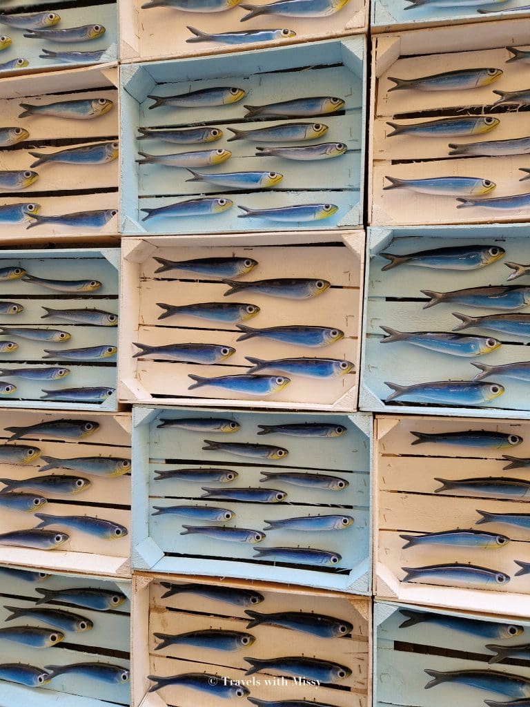 Boxes of ceramic sardines are displayed in wooden boxes and 6 blue sardines are in each box. One of many
