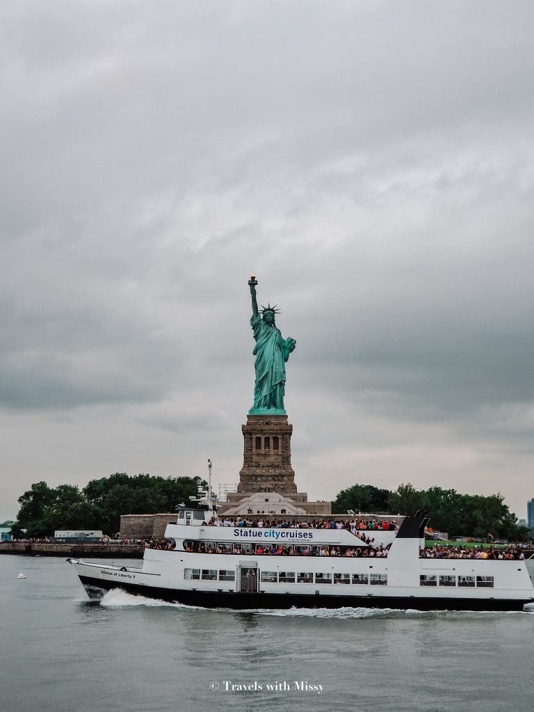 The Statue of Liberty poses in front of a Statue City Cruises which sails past on a gloomy summer day. 