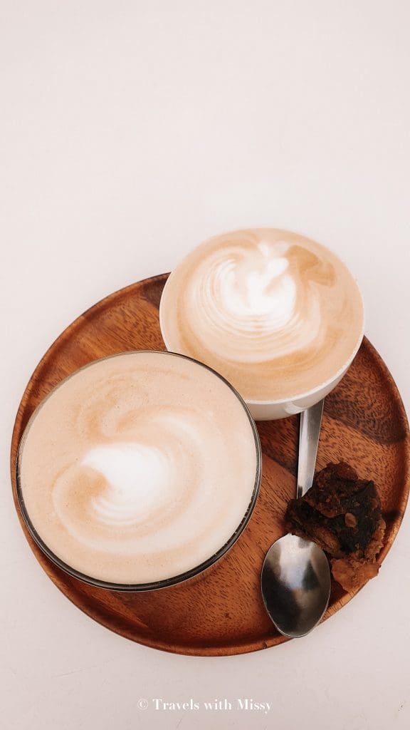 Two cups of coffee placed on a brown circular tray with a small piece of cake.